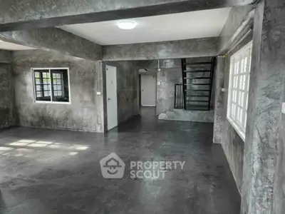 Spacious industrial-style living room with concrete walls and floors, featuring large windows and a staircase.