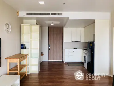 Modern studio apartment with open layout kitchen featuring white cabinetry and built-in appliances, complemented by rich wooden flooring and ample natural light.