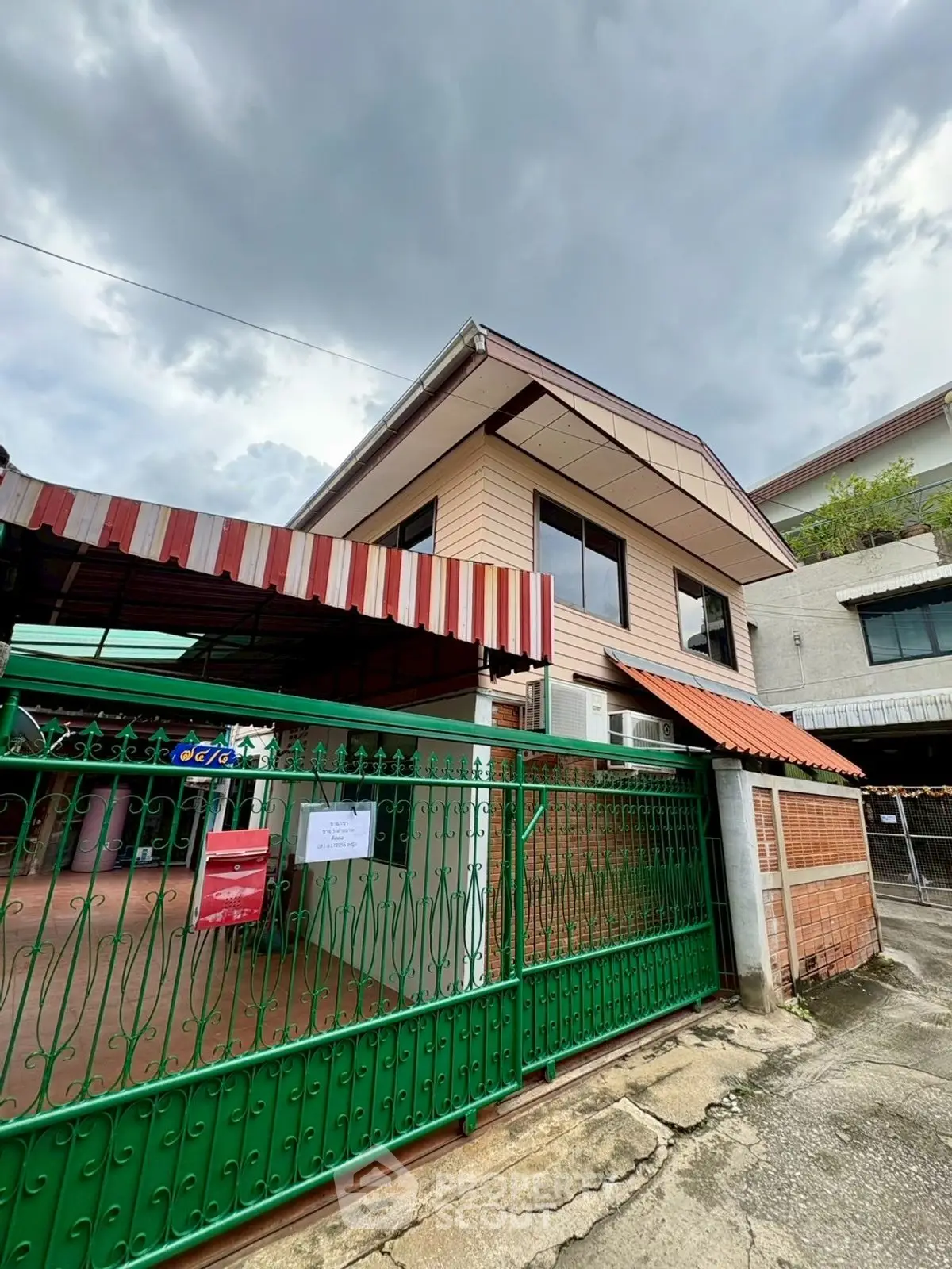 Charming two-story house with green gate and covered carport under cloudy skies.