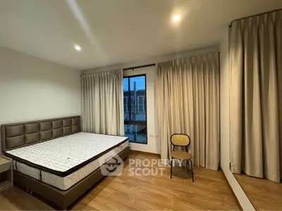 Spacious bedroom with wooden flooring and large window, featuring a comfortable bed and elegant curtains.