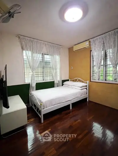 Cozy bedroom with wooden flooring, white metal bed, and large windows with curtains.
