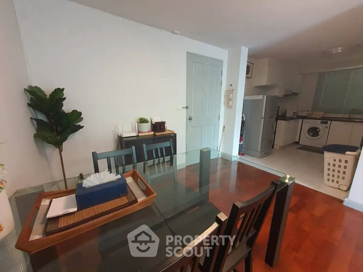Modern dining area with glass table, wooden chairs, and adjacent kitchen featuring appliances.