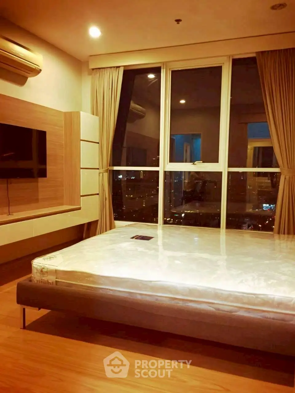 Modern bedroom with large window and city view, featuring a mounted TV and air conditioning.
