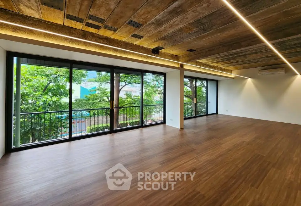 Spacious modern living room with large windows and wooden flooring, offering a serene view of lush greenery.