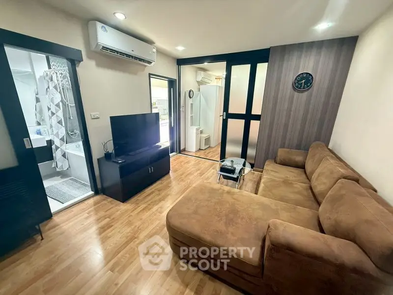 Modern living room with brown sectional sofa and wall-mounted TV, adjacent to a bathroom.