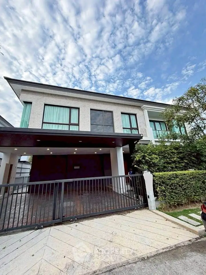 Modern two-story house with large windows and gated driveway, surrounded by lush greenery.