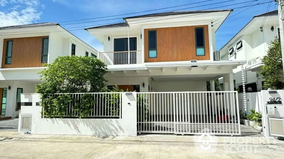 Modern two-story house with white facade and wooden accents, featuring a gated entrance and lush greenery.