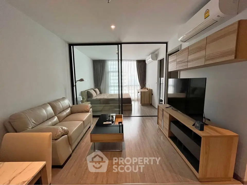 Modern open-plan living room with glass partition leading to a stylish bedroom, featuring sleek furniture and natural light.