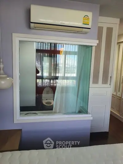Modern bedroom with unique glass partition overlooking bathroom, featuring built-in wardrobe and air conditioning.