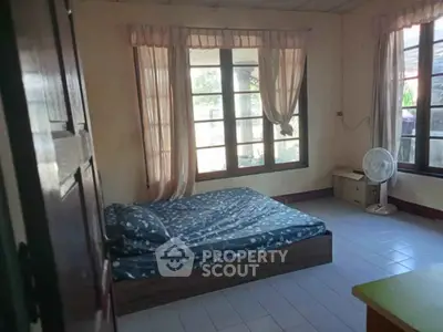 Cozy bedroom with large windows and natural light, featuring a simple bed and fan.